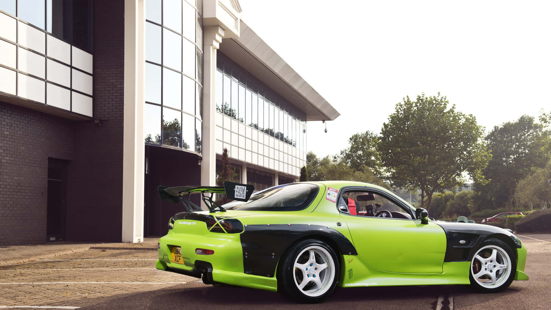  壁紙 — 一輛充滿活力的萊姆綠色和黑色馬自達RX-7跑車，帶有大型後尾翼，停在一棟現代玻璃建築外。
