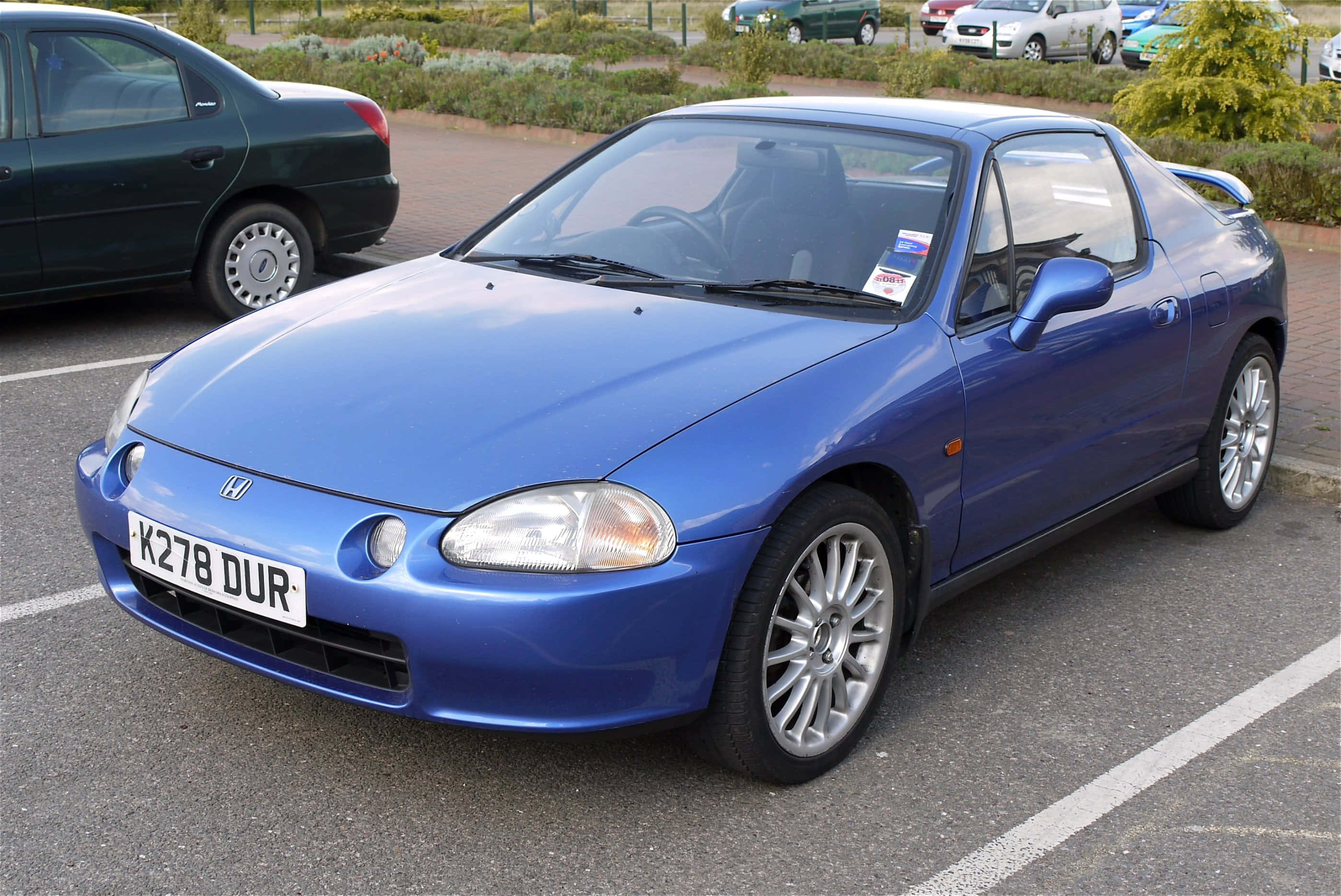 Honda Civic Del Sol 桌布 壁紙 — 一輛充滿活力的藍色本田 Del Sol 敞篷車停放在日間停車場，背景有其他車輛和綠化。