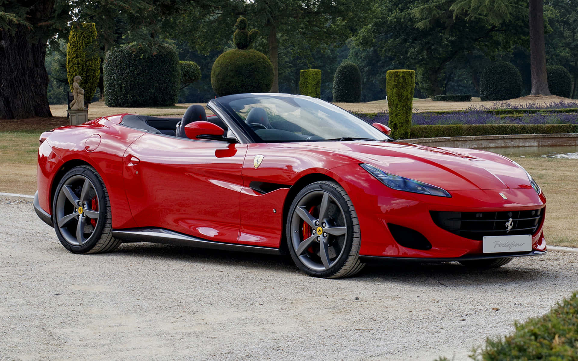 Ferrari Portofino 桌布 壁紙 — 一輛鮮紅色的法拉利 Portofino 敞篷車在日光下閃耀，行駛在由修剪整齊的花園和雕塑式樹籬環繞的碎石路上。