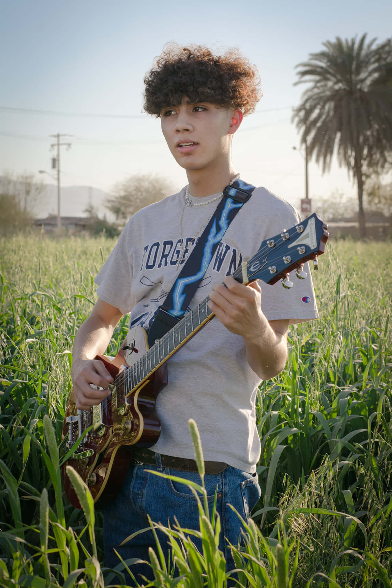  壁紙 — 一位捲髮年輕男子在陽光明媚的高大綠草田野中彈奏紅色電吉他，背景有棕櫚樹。