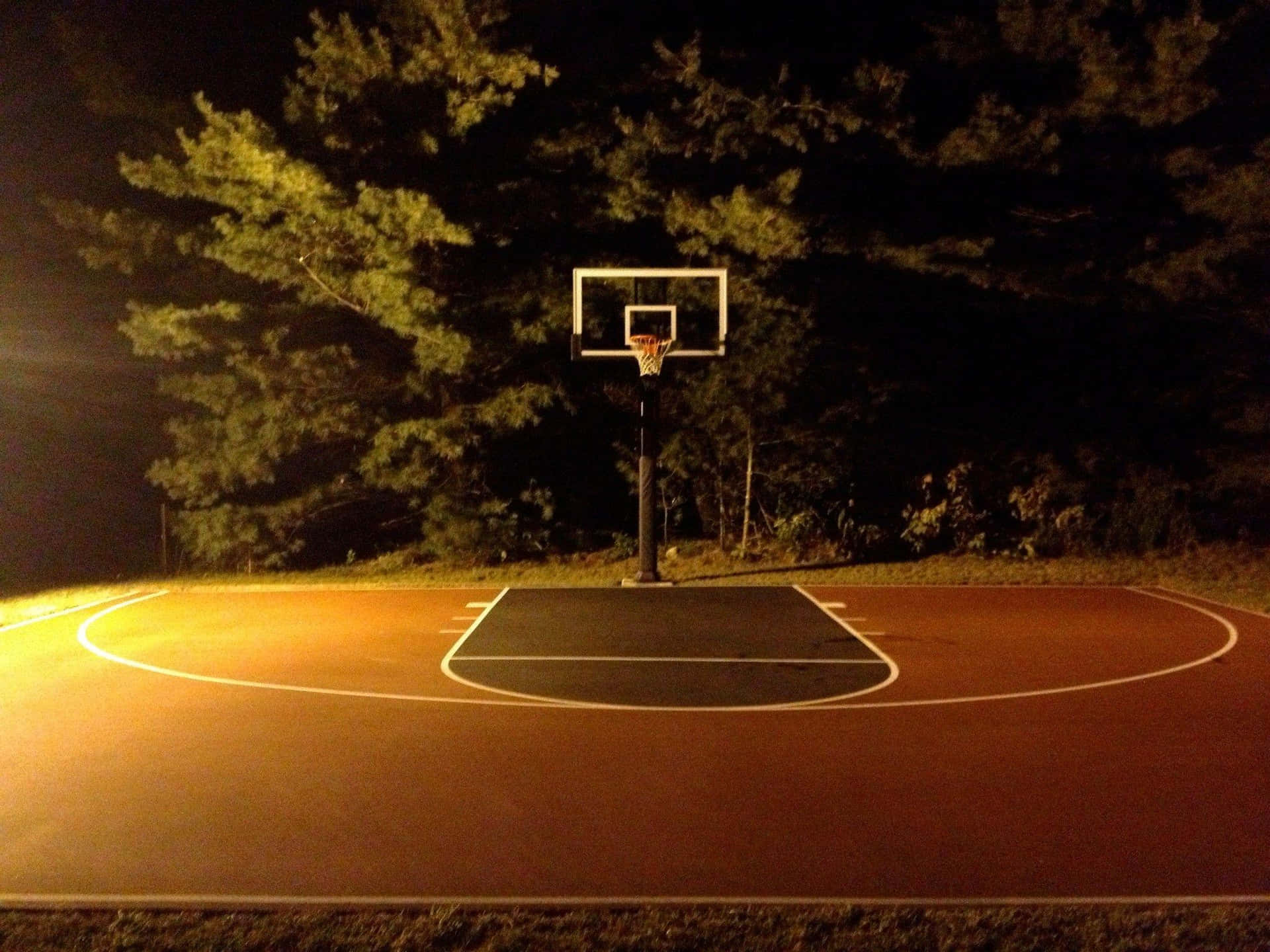  壁紙 — 夜晚被溫暖人造光照亮的籃球場，背景有樹木剪影。
