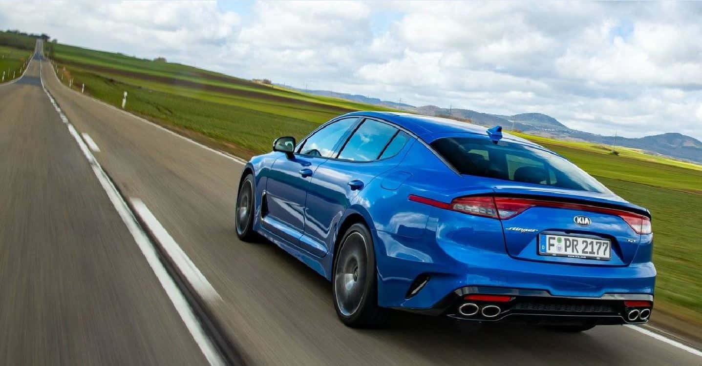 Kia Stinger 桌布 壁紙 — 一輛充滿活力的藍色Kia Stinger GT在鄉間道路上疾馳，背景是模糊的綠色田野和遠處的山丘，天空多雲。