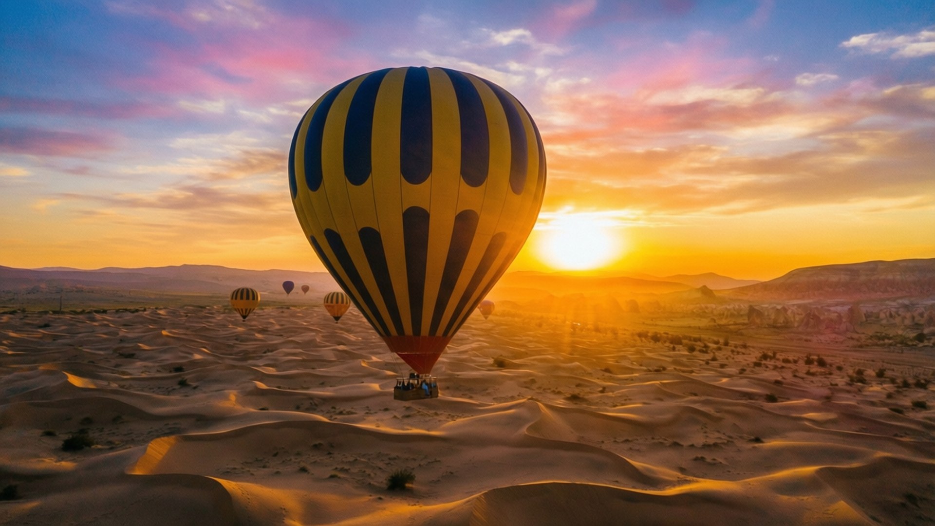Balloon Ride Sunset 壁紙 — 一個黃藍相間的熱氣球在日落時分飄浮在金黃色的沙丘上空，遠處還有其他氣球。