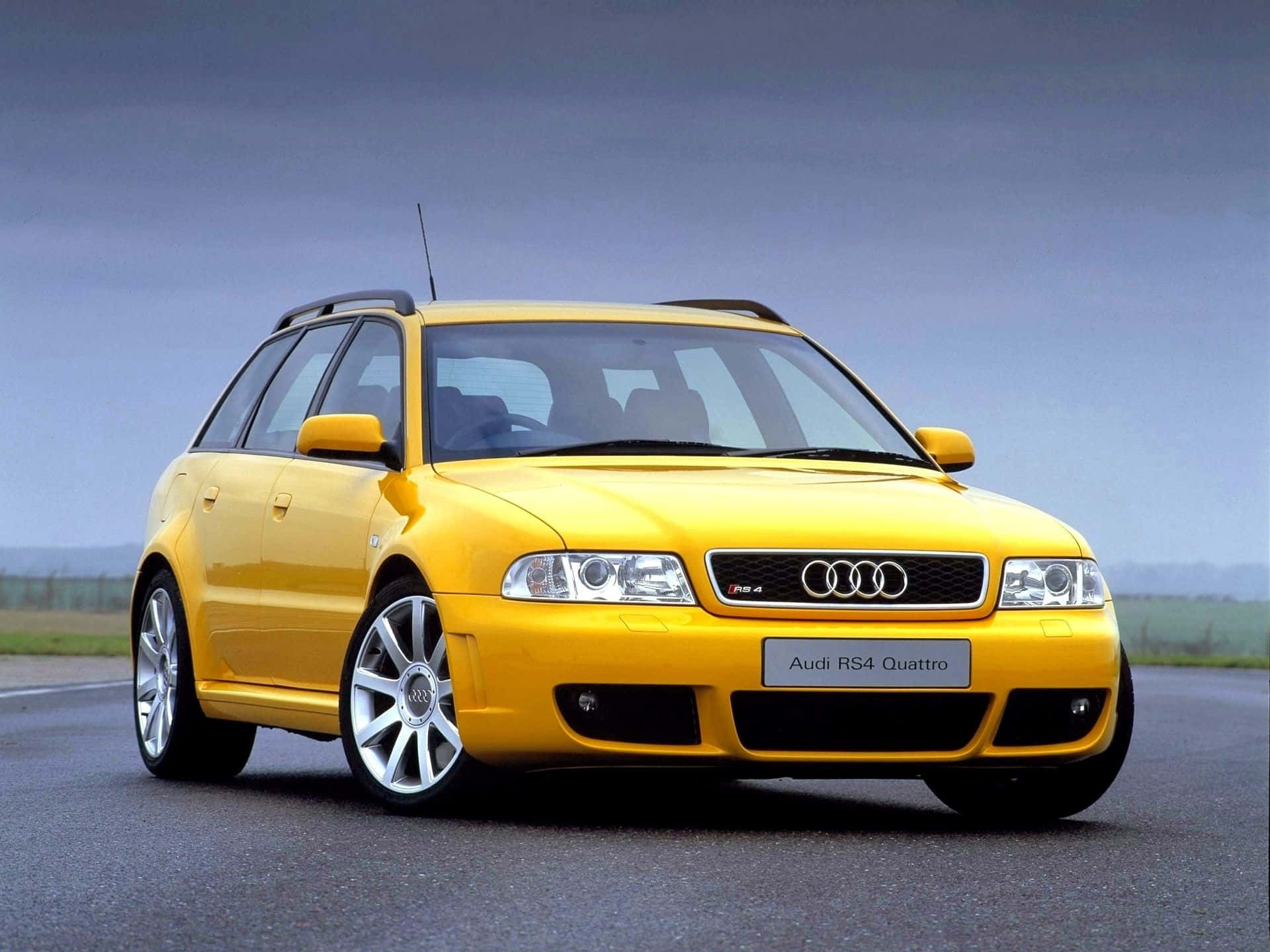 Audi Rs4 桌布 壁紙 — 一輛鮮黃色的奧迪RS4 Quattro旅行車停放在柏油路上，天空多雲。