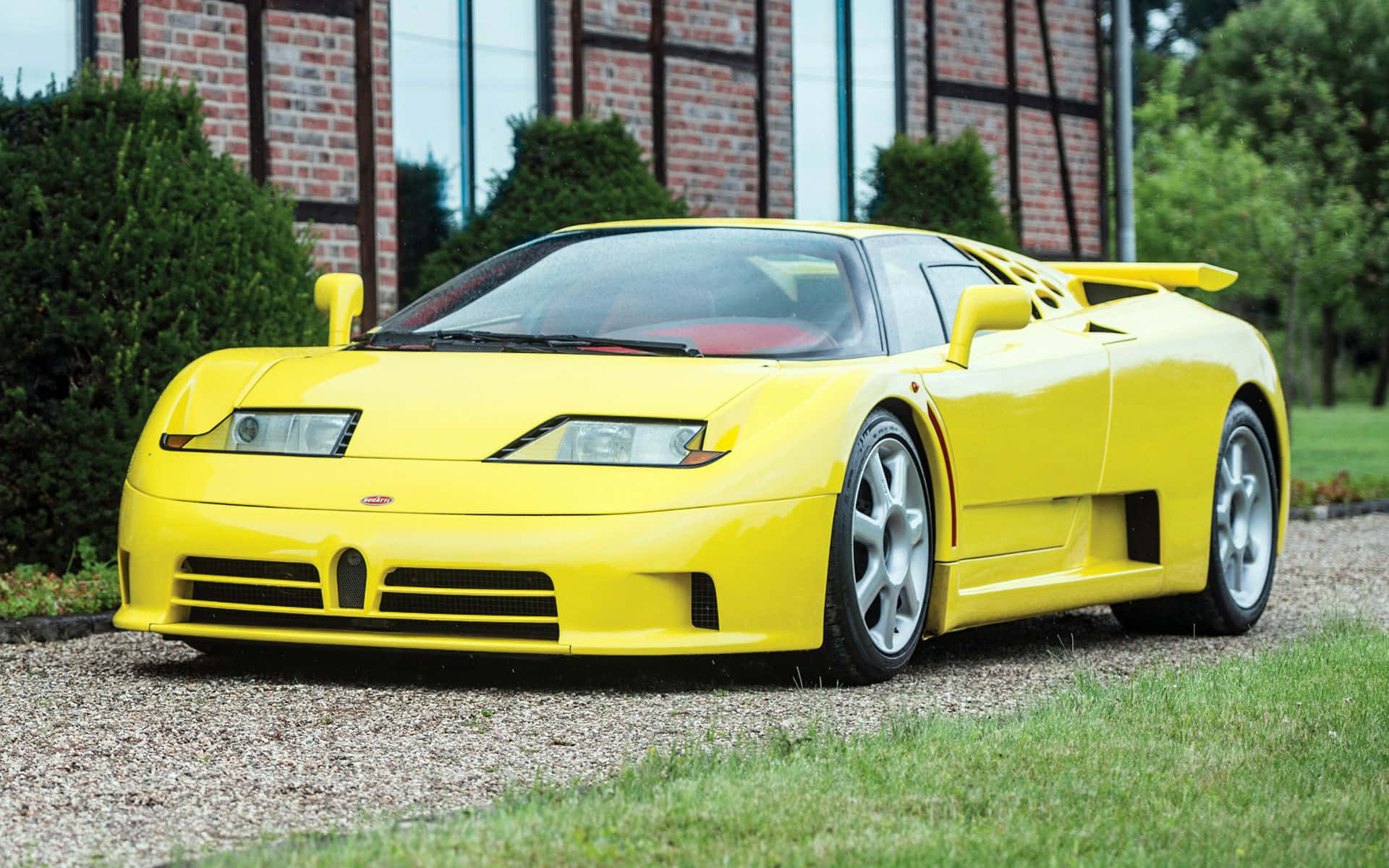 Bugatti Eb110 桌布 壁紙 — 一輛鮮黃色的Bugatti EB110超級跑車停放在磚造建築前的碎石地上，周圍有綠植。