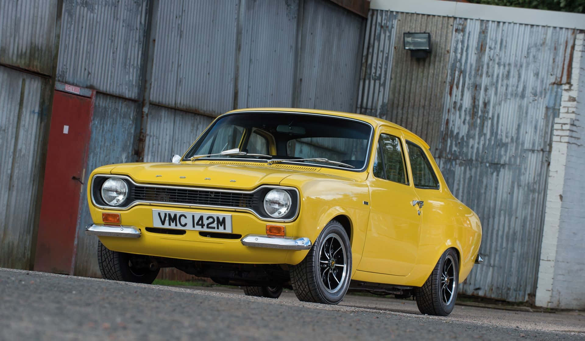 Ford Escort 桌布 壁紙 — 一輛鮮黃色的經典福特福克斯停泊在風化的波紋金屬建築前。
