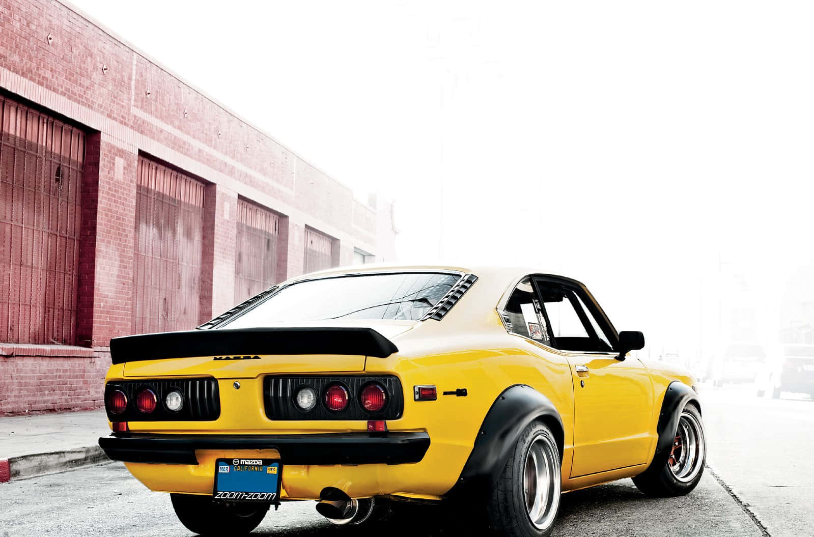 Mazda Rx-3 桌布 壁紙 — 一輛鮮黃色經典馬自達RX-7跑車，帶黑色點綴，停在磚砌倉庫旁，在明亮日光下。