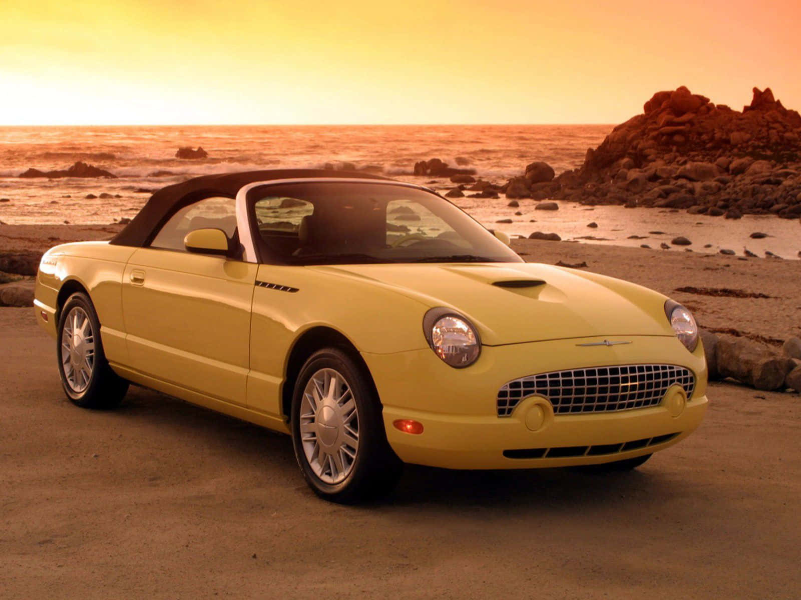Ford Thunderbird 桌布 壁紙 — 一輛黃色敞篷跑車停泊在岩石海灘上，夕陽西下，溫暖的橙色光線反射在海面上。