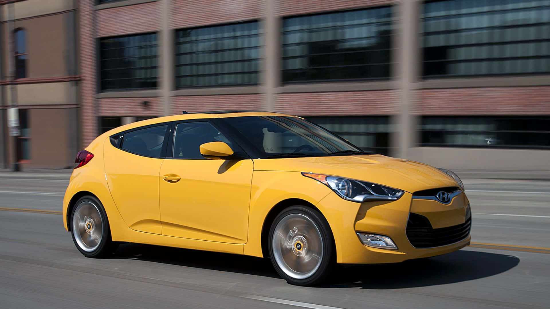 Hyundai Veloster 桌布 壁紙 — 一輛充滿活力的黃色現代Veloster在磚砌建築背景下，以動態模糊效果在城市街道上飛馳。