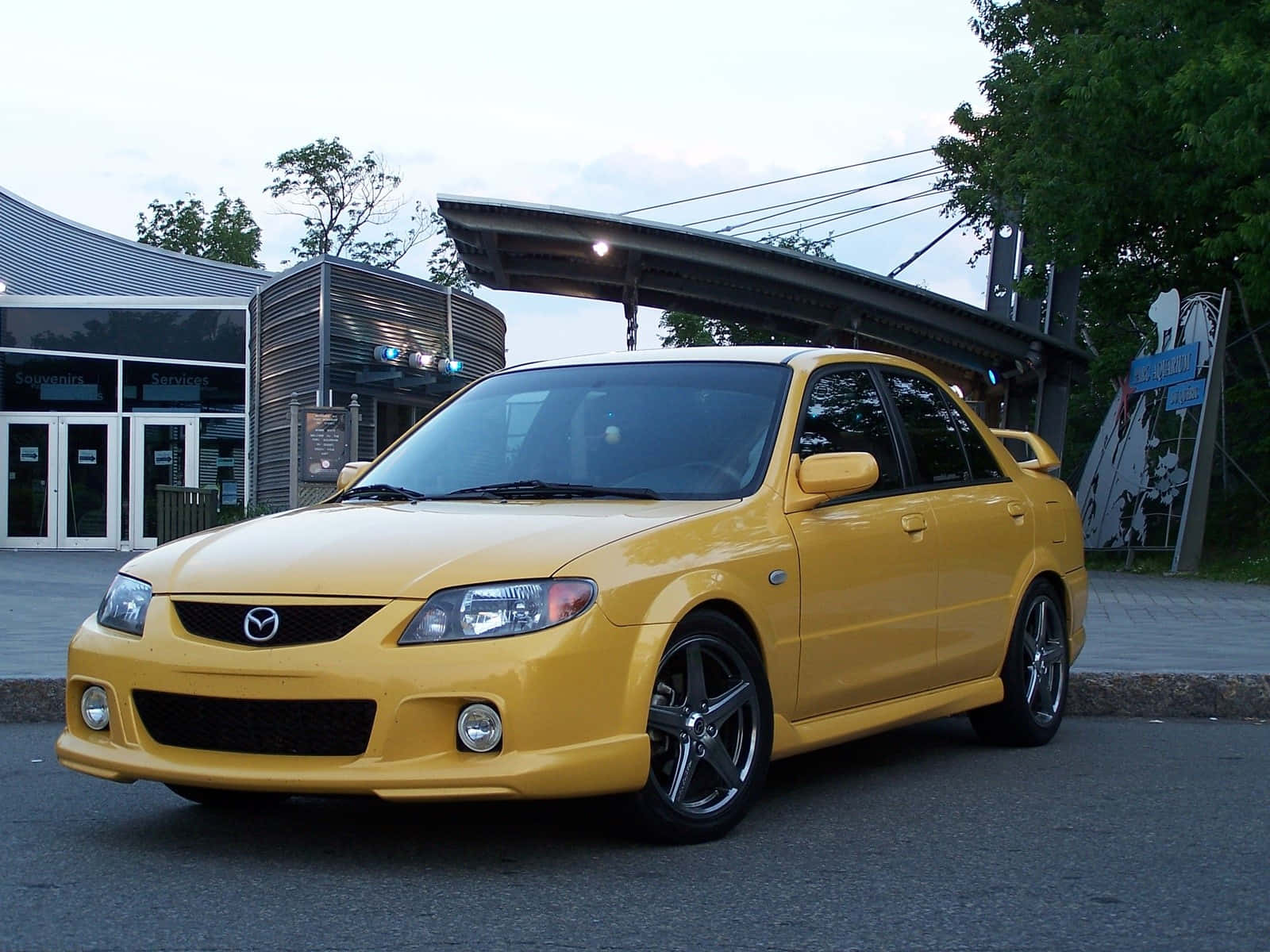 Mazda Protege 桌布 壁紙 — 一輛鮮黃色的Mazda 3轎車，配黑色輪圈，在黃昏時停泊在現代建築附近的柏油路上。