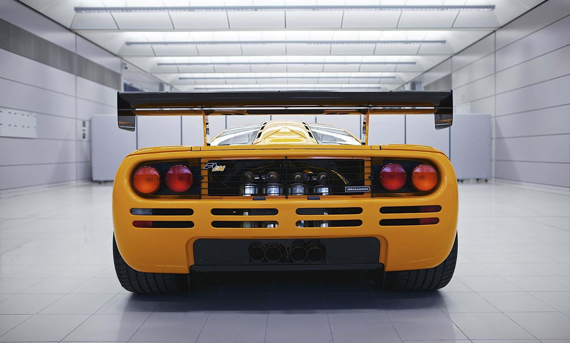Mclaren F1 桌布 壁紙 — 一輛充滿活力的黃色McLaren F1超級跑車從後方觀看，位於明亮無菌的車庫中，帶有大型後翼和暴露的引擎。