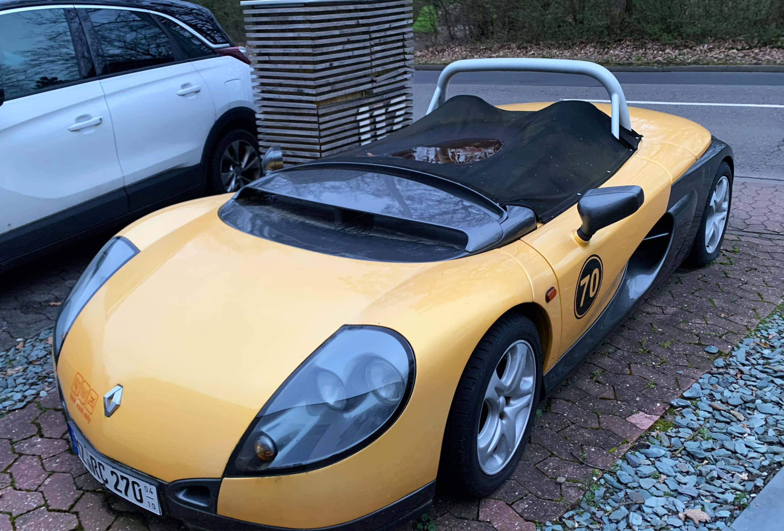 Renault Sport Spider 桌布 壁紙 — 一輛亮黃色Renault Spider跑車，帶有黑色飾邊和70號碼，停泊在鵝卵石街道上，旁邊是一輛白色SUV。