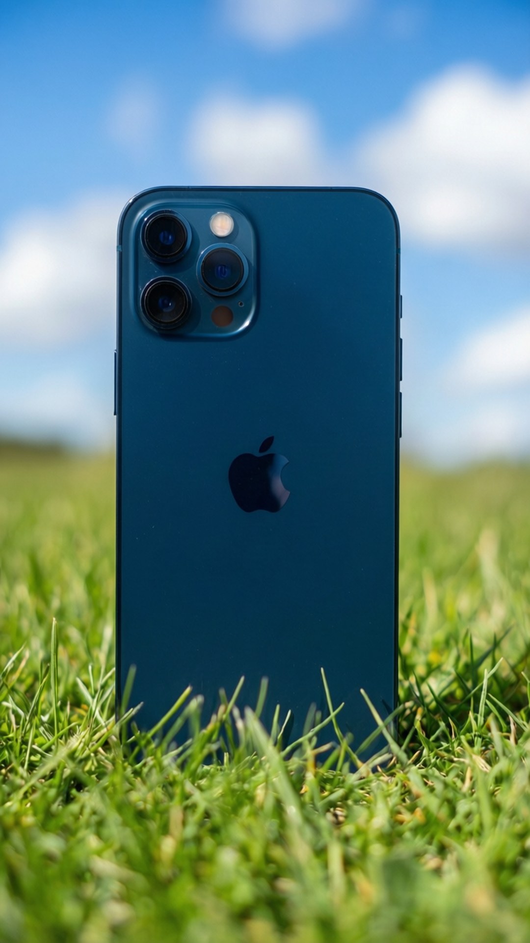 Sky Blue Harmony 壁紙 — 一部藍色iPhone配備三鏡頭相機，直立於充滿活力的綠色草地上，在明亮的藍天下有蓬鬆的白雲。