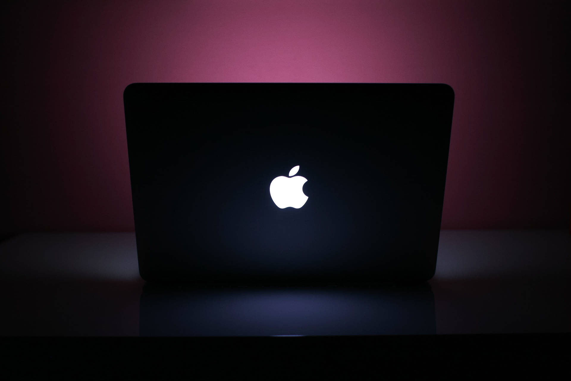 黑色筆記型電腦，帶有發光的Apple標誌，在粉紅色燈光下的深色表面上。 — Macbook Pro 壁紙