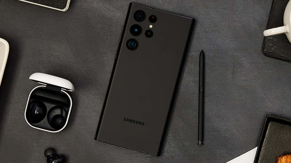  壁紙 — 一部時尚的黑色Samsung智慧型手機置於深灰色紋理表面中央，周圍有S Pen、耳機和一個白色咖啡杯。