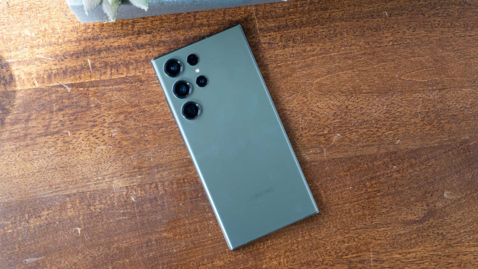  壁紙 — 一部時尚的鼠尾草綠色Samsung智慧型手機以對角線方式放置在溫暖有紋理的木質表面上，伴隨著柔和自然光。