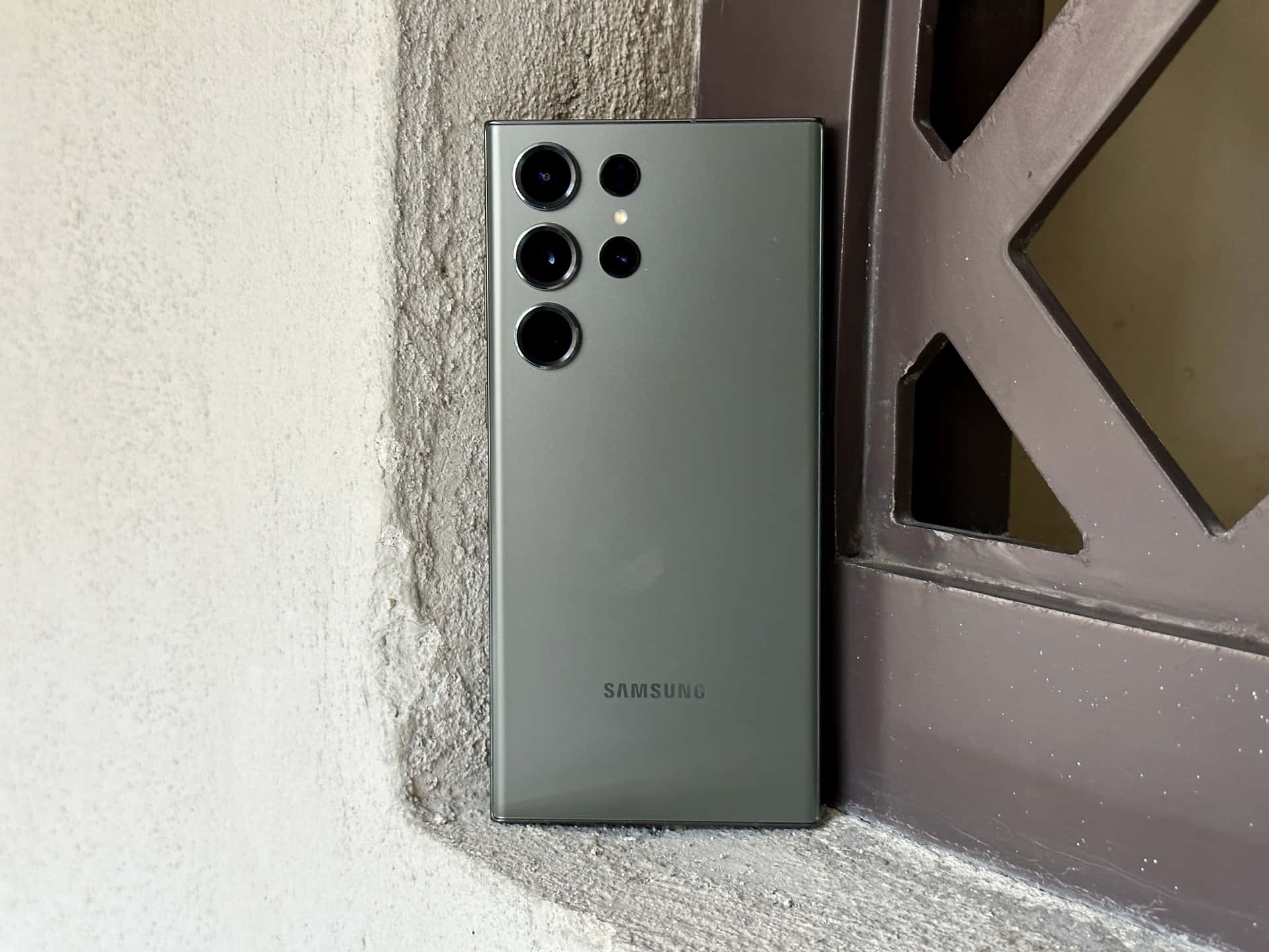 壁紙 — 一部時尚的霧面綠色Samsung智慧型手機靠在一面有紋理的牆上，旁邊是一扇幾何金屬門。