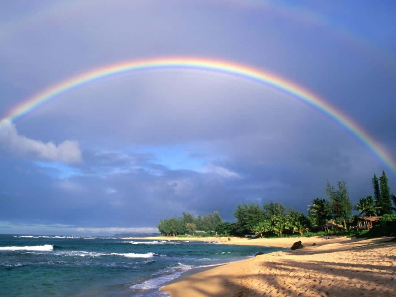  壁紙 — 一道鮮豔的彩虹在熱帶海灘上空拱起，碧綠的海浪拍打著金色沙灘，天空多雲。