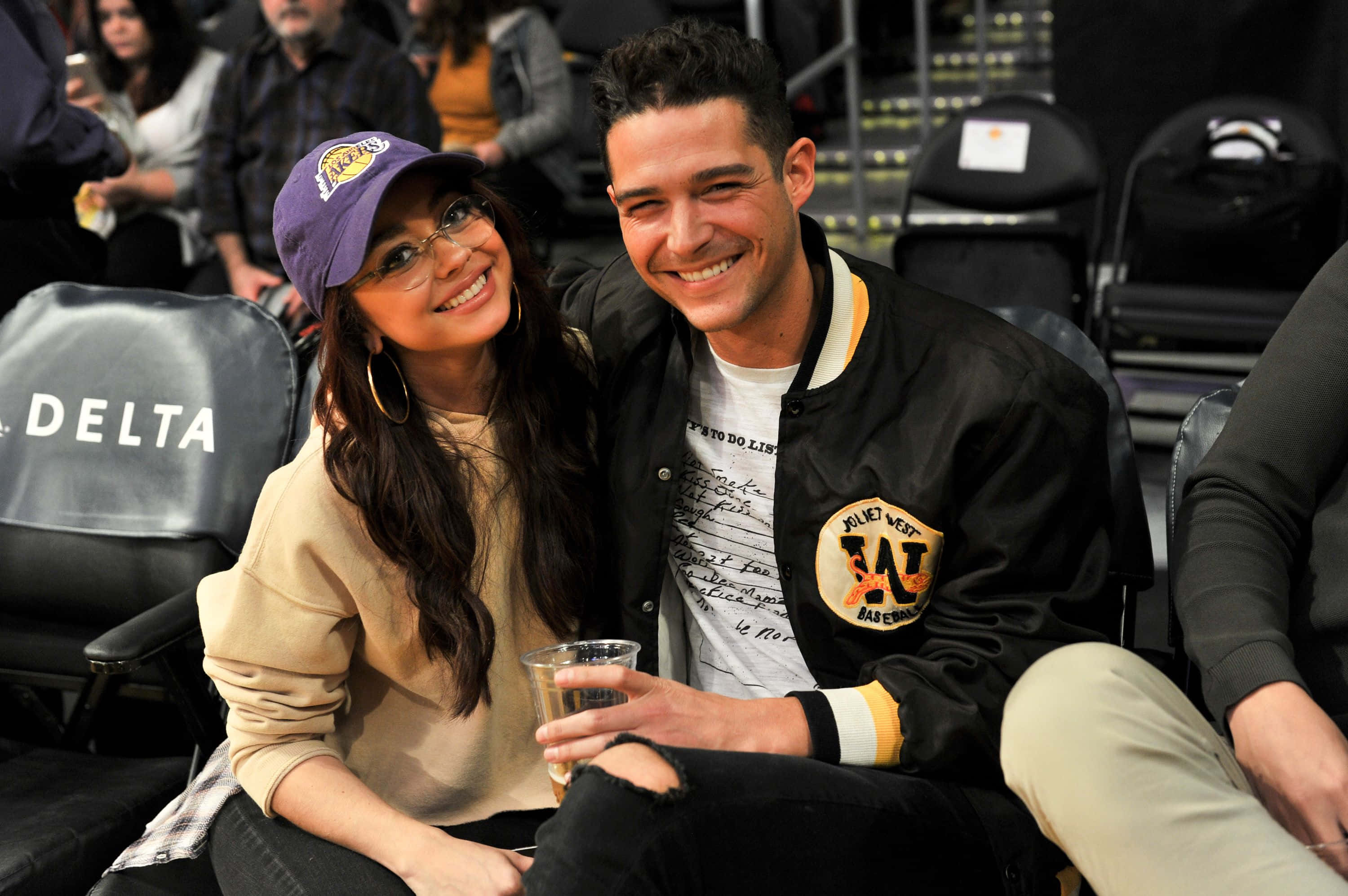 Sarah Hyland 桌布 壁紙 — 一對微笑的情侶在籃球場上，女士戴著紫色湖人隊帽子，男士穿著黑色飛行員夾克拿著飲料。