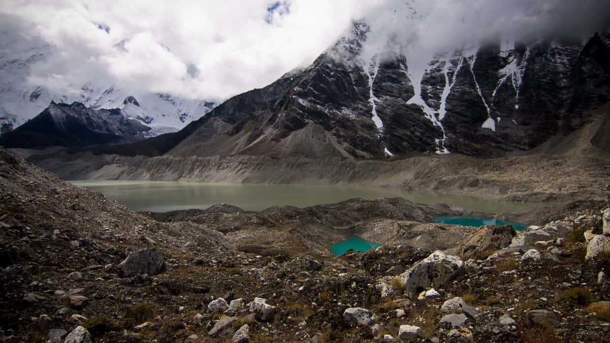  壁紙 — 一個高海拔的高山湖泊，坐落在積雪的山峰和岩石斜坡之間，天空多雲。