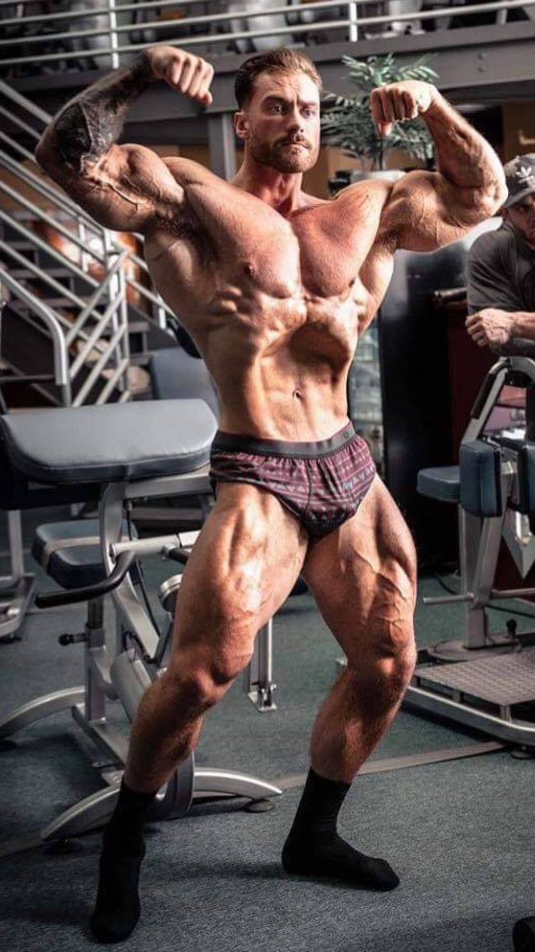 一個肌肉男在健身房中繃緊手臂。 — Chris Bumstead 壁紙