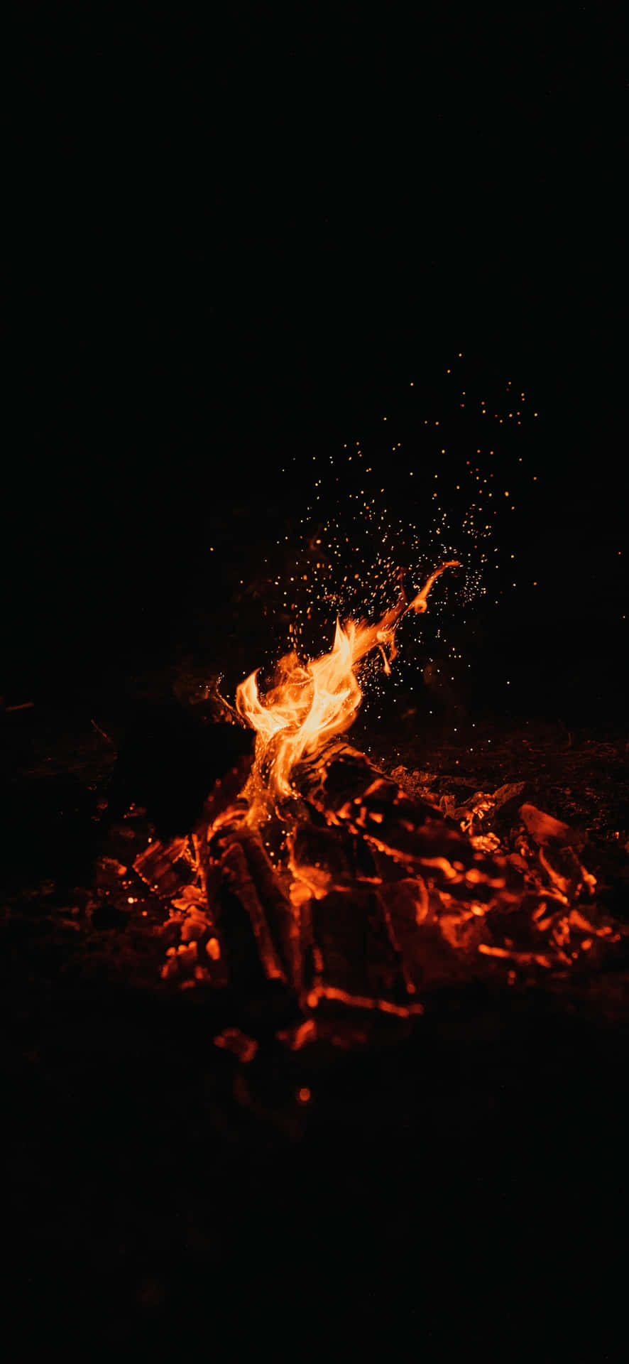  壁紙 — 一個充滿活力的營火在漆黑的夜空中劈啪作響並迸出火花，將溫暖的橙色光芒投射在發光的餘燼上。