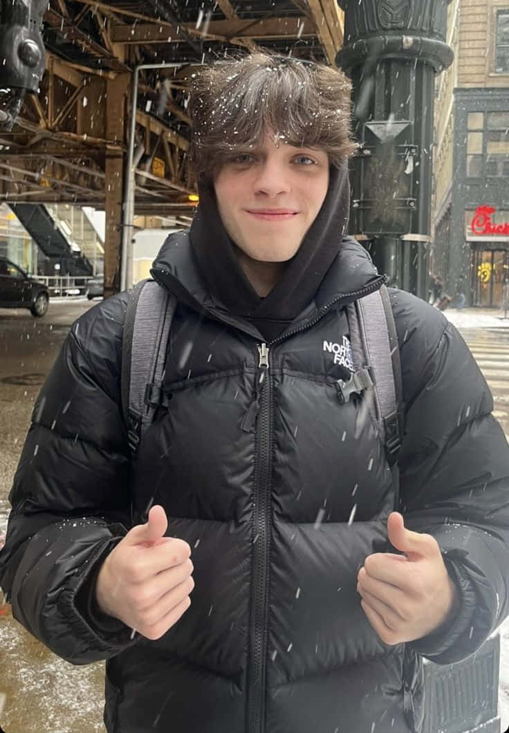  壁紙 — 一個穿著黑色羽絨外套的年輕男子站在城市街道上飄落的雪中，豎起兩個大拇指。