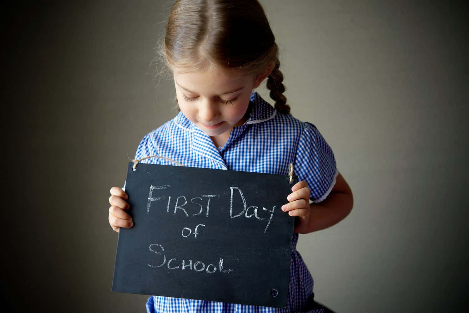  壁紙 — 一個穿著藍色格子校服的年輕女孩，專注地拿著一塊寫著「First Day of School」的黑板標語牌。