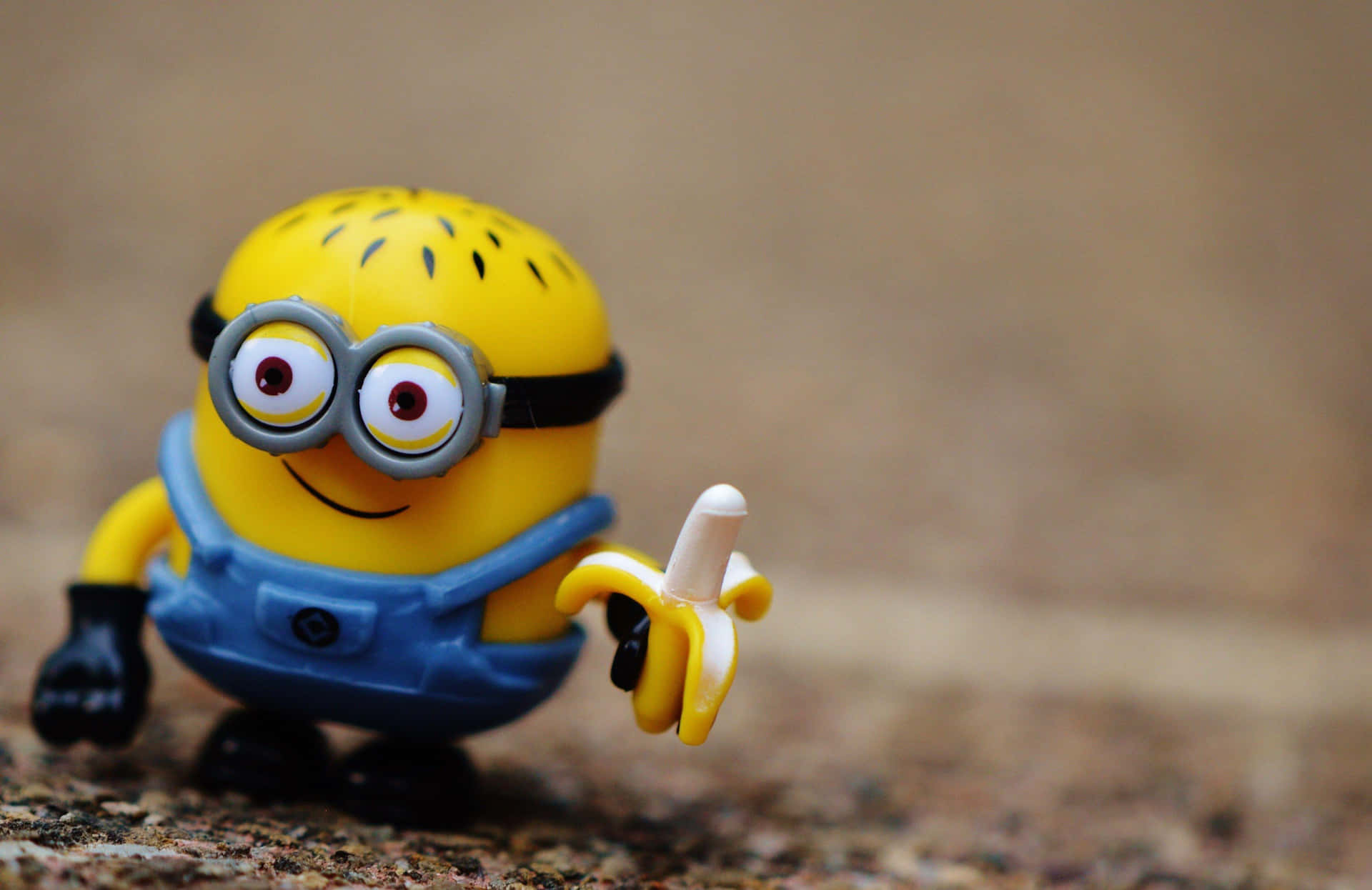  壁紙 — 一個戴著護目鏡、開心的黃色Minion玩具，在模糊的泥土背景前拿著一根香蕉。