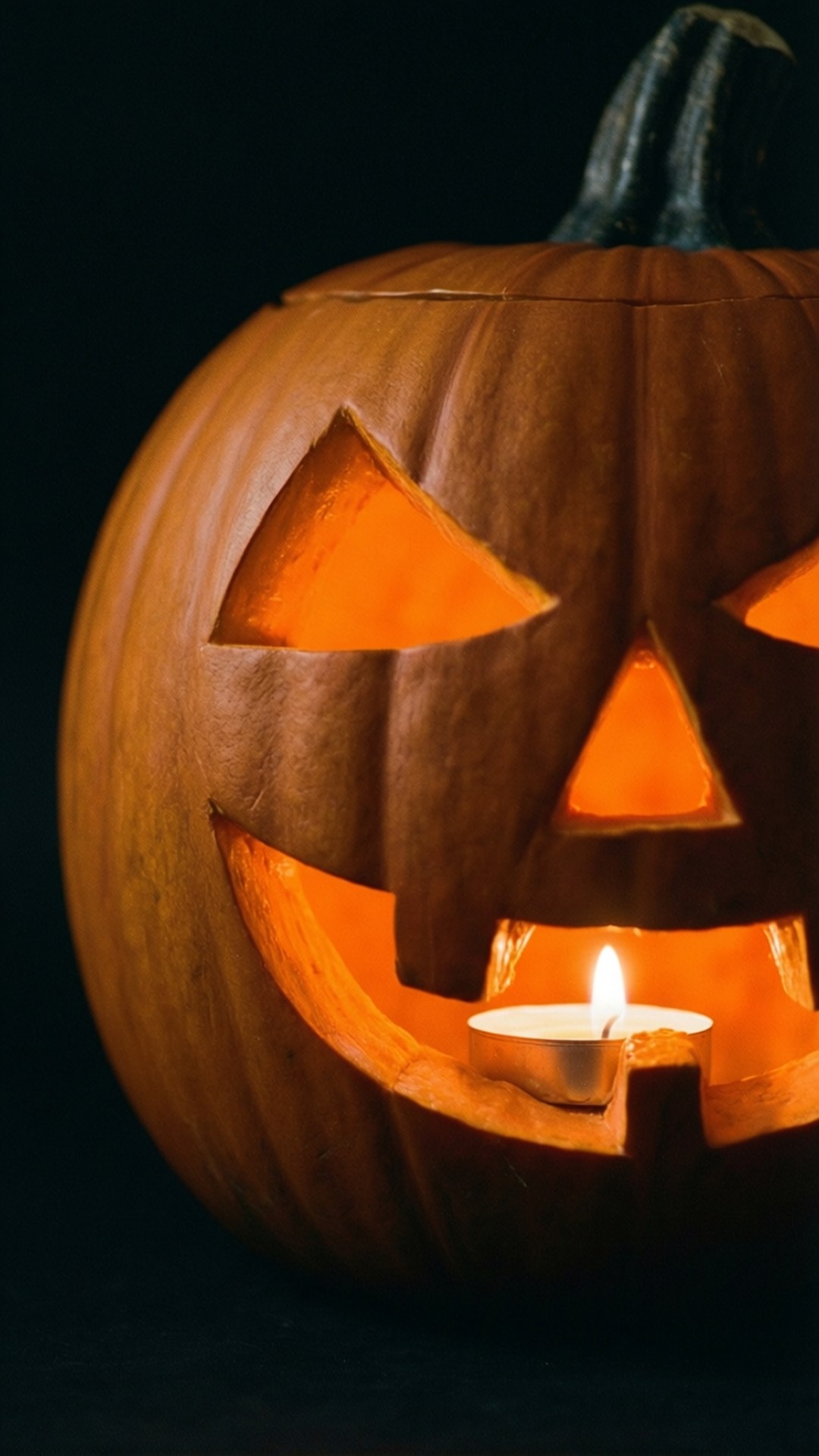 Glowing Jack-O'-Lantern 壁紙 — 一個雕刻的南瓜，有三角形眼睛和寬闊的笑容，內裡的蠟燭在黑色背景下溫暖地發光。