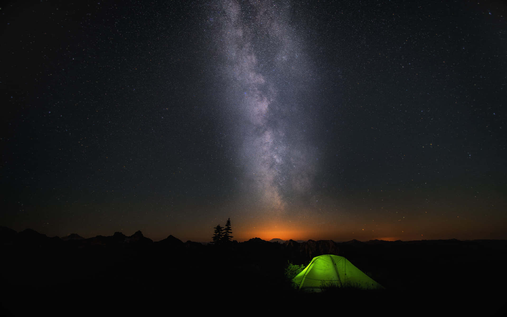 一個發光的綠色帳篷在星空下，銀河在山脈上方。 — Windows 11 壁紙