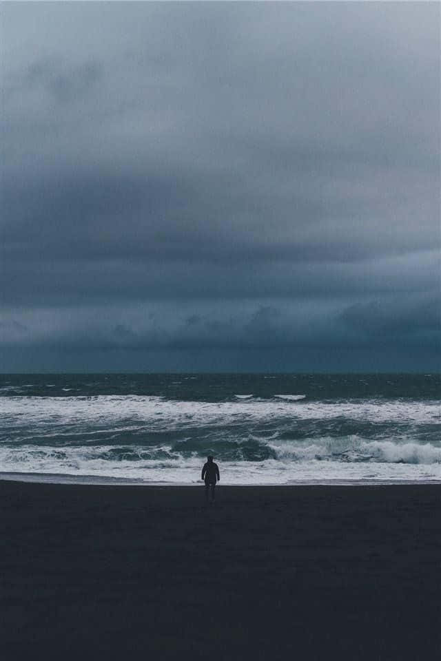  壁紙 — 一個孤獨的身影沿著黑暗的海灘行走，暴風雨般的海浪在沉重的灰色天空下拍打。
