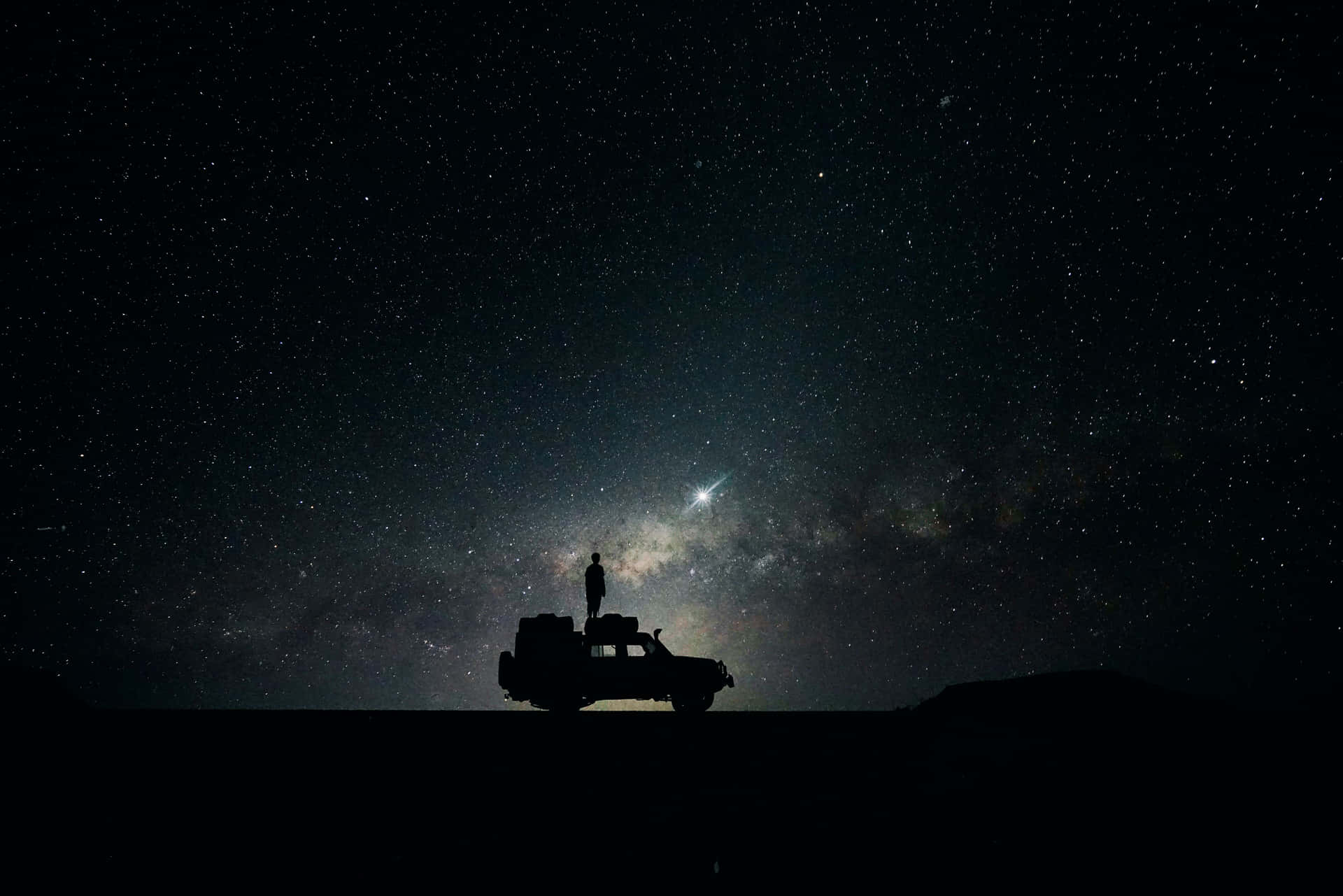  壁紙 — 一個孤獨的身影站在堅固的車輛上，在令人嘆為觀止的星空夜空下，銀河在上方閃耀。