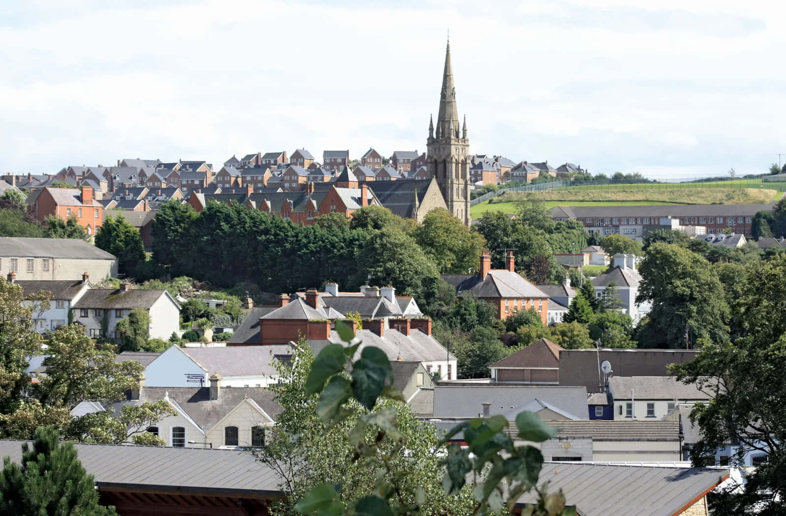 Newry United Kingdom 桌布 壁紙 — 一個古色古香的小鎮全景，擁有紅磚房屋和高聳的教堂尖塔，在陰雲密布的天空下，聳立在茂密的綠樹之上。