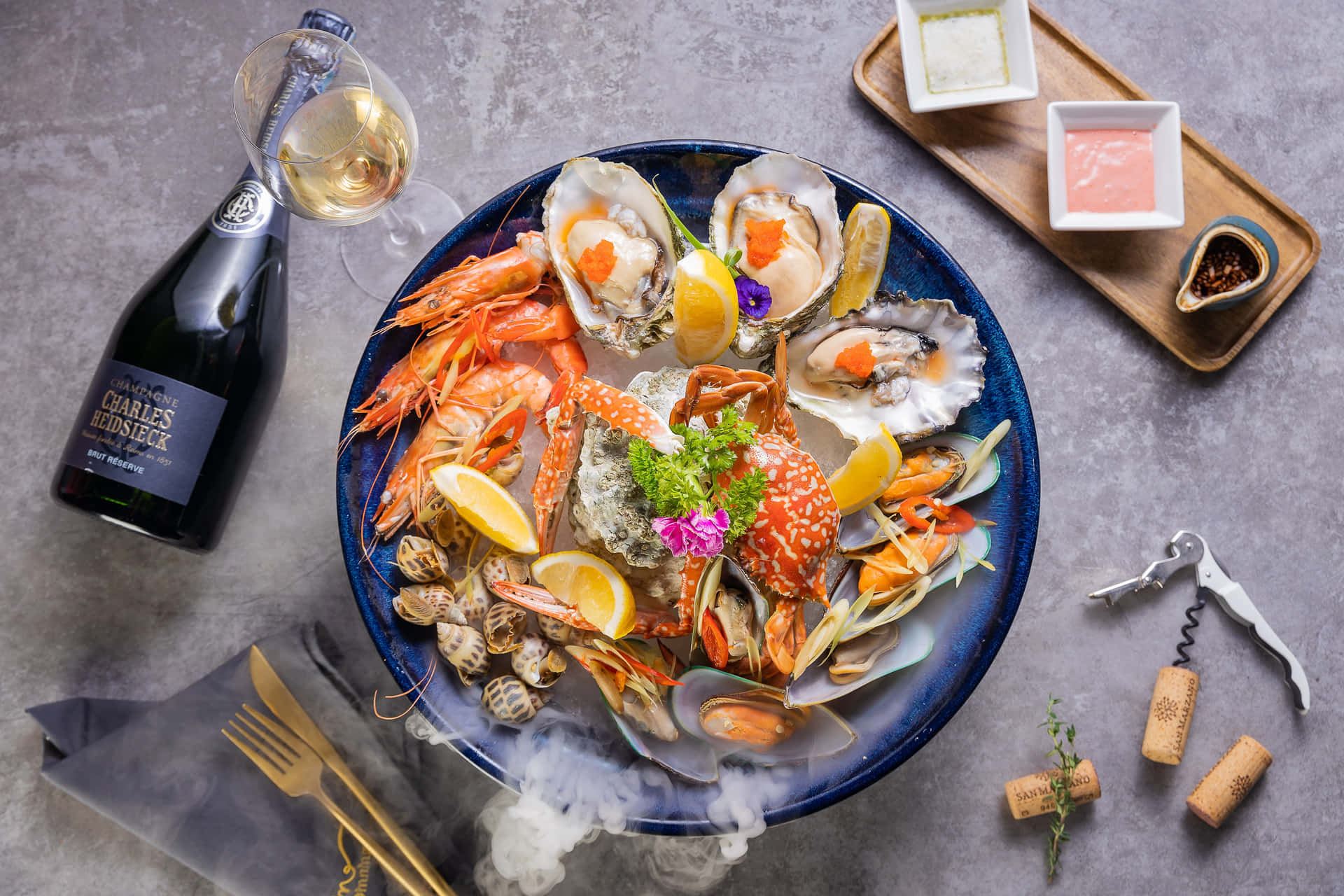  壁紙 — 一個豪華海鮮拼盤，有生蠔、蝦、蟹和淡菜，以檸檬和香草裝飾，旁邊放著一瓶香檳和軟木塞，在灰色石面上。
