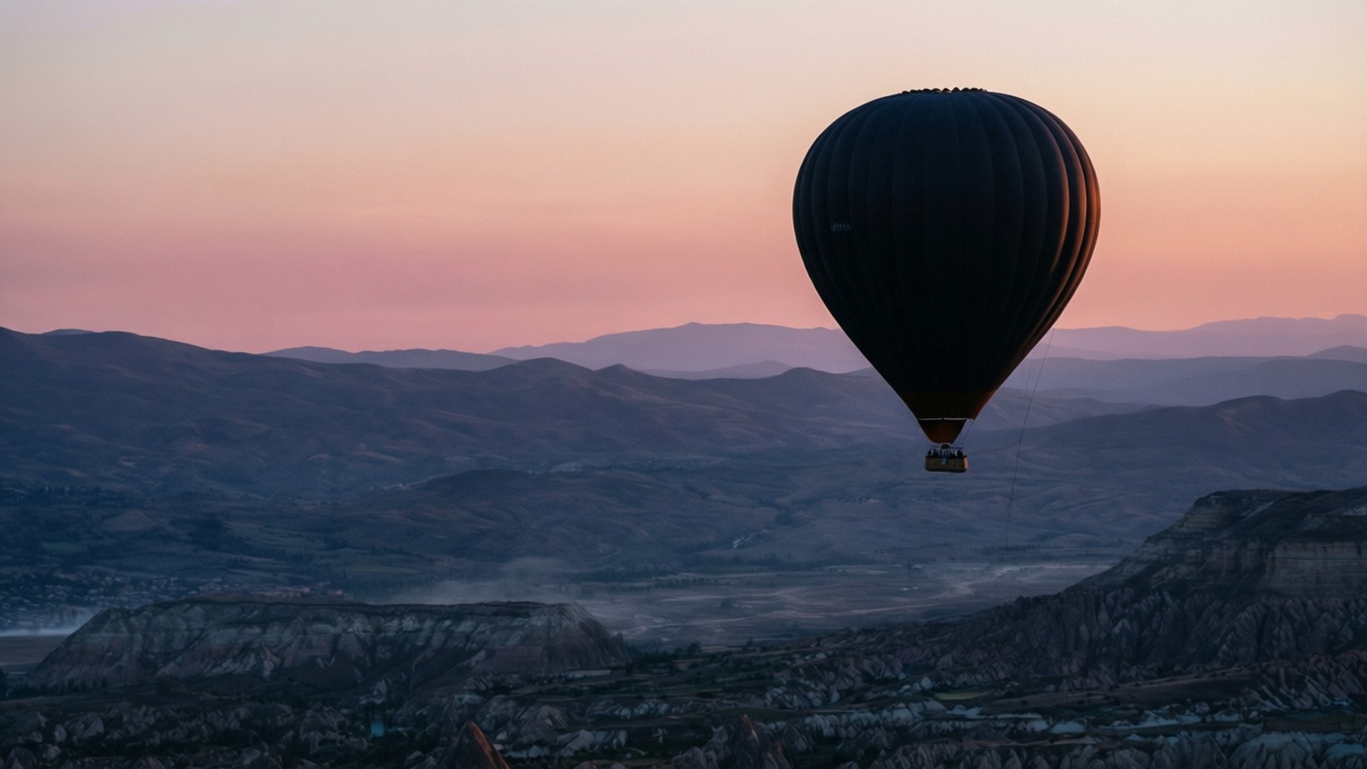 Balloon Over Valley Silhouette 壁紙 — 一個黑色熱氣球在暮色中平靜地飄浮在崎嶇的山脈之上，伴隨著柔和的粉紅色天空。