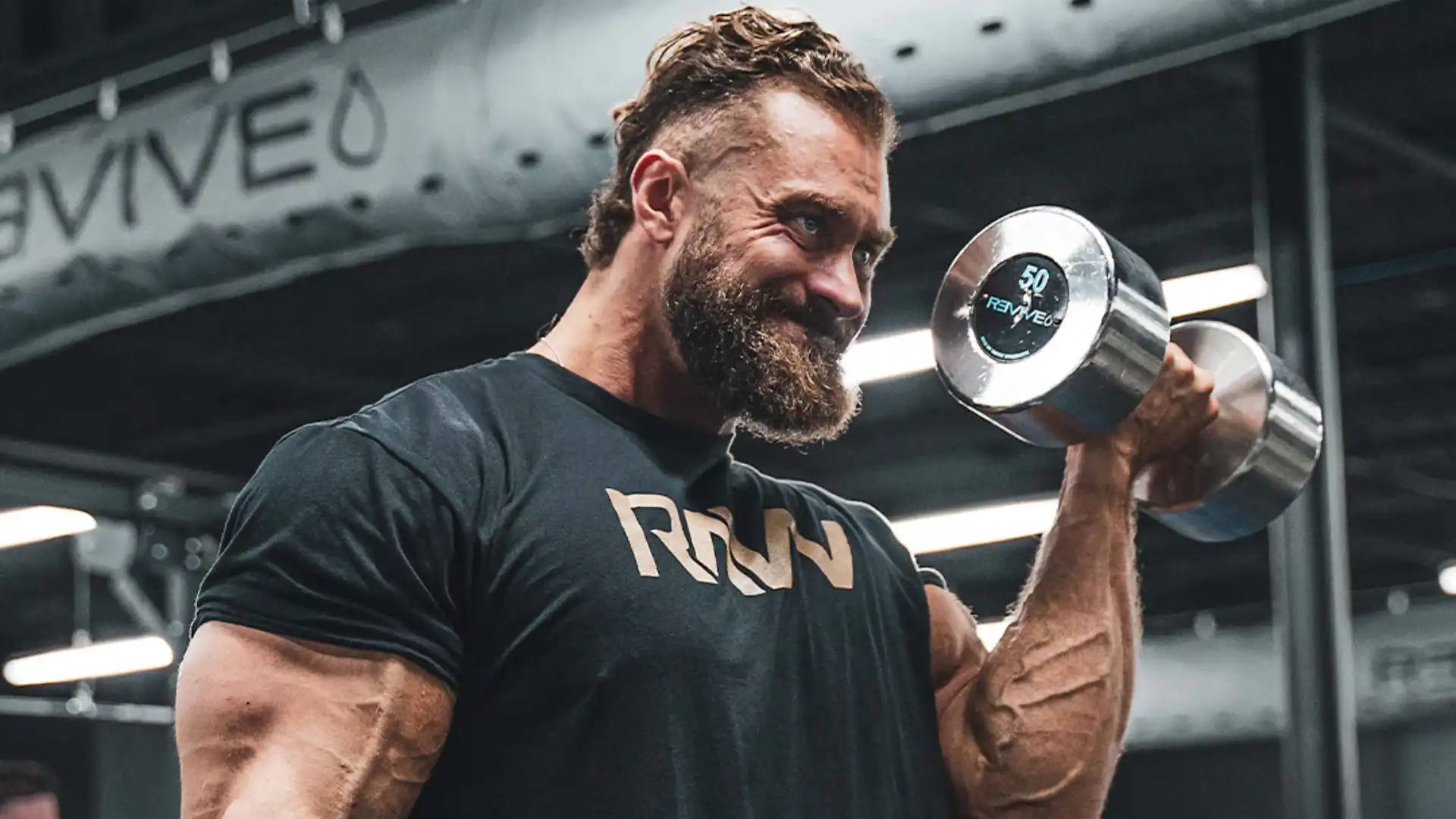 鬍鬚男在工業照明健身房舉起50磅啞鈴 — Chris Bumstead 壁紙