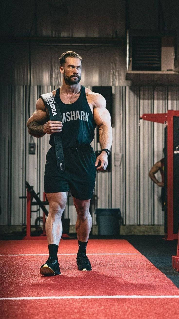 一個肌肉發達的男人在健身房拿著舉重腰帶。 — Chris Bumstead 壁紙