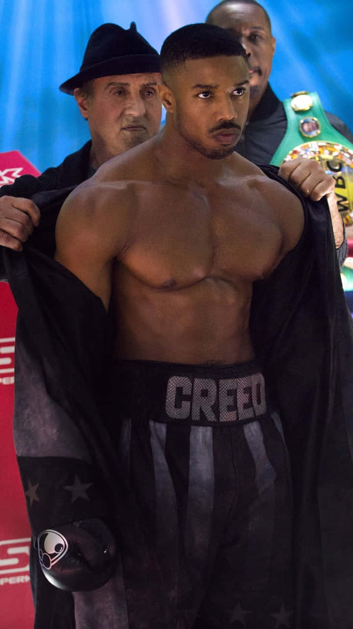  壁紙 — 一個肌肉發達的拳擊手穿著黑色 'CREED' 短褲，赤裸上身，一個戴著軟呢帽的男人拉開他的長袍，露出他結實的身材。