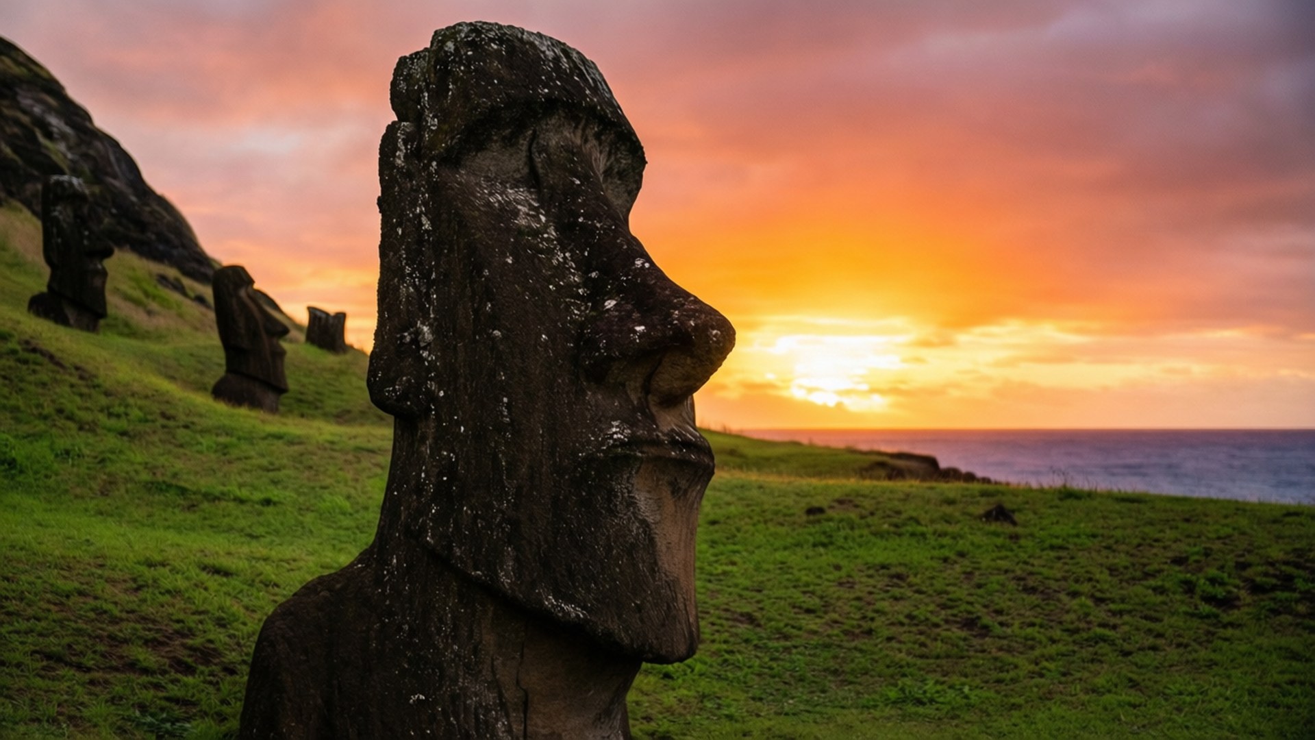 Moai Statue Sunset Silhouette 壁紙 — 一個巨大的石頭摩艾雕像在日落時分，以橙色天空和海洋為背景，側面站在長滿草的山丘上。
