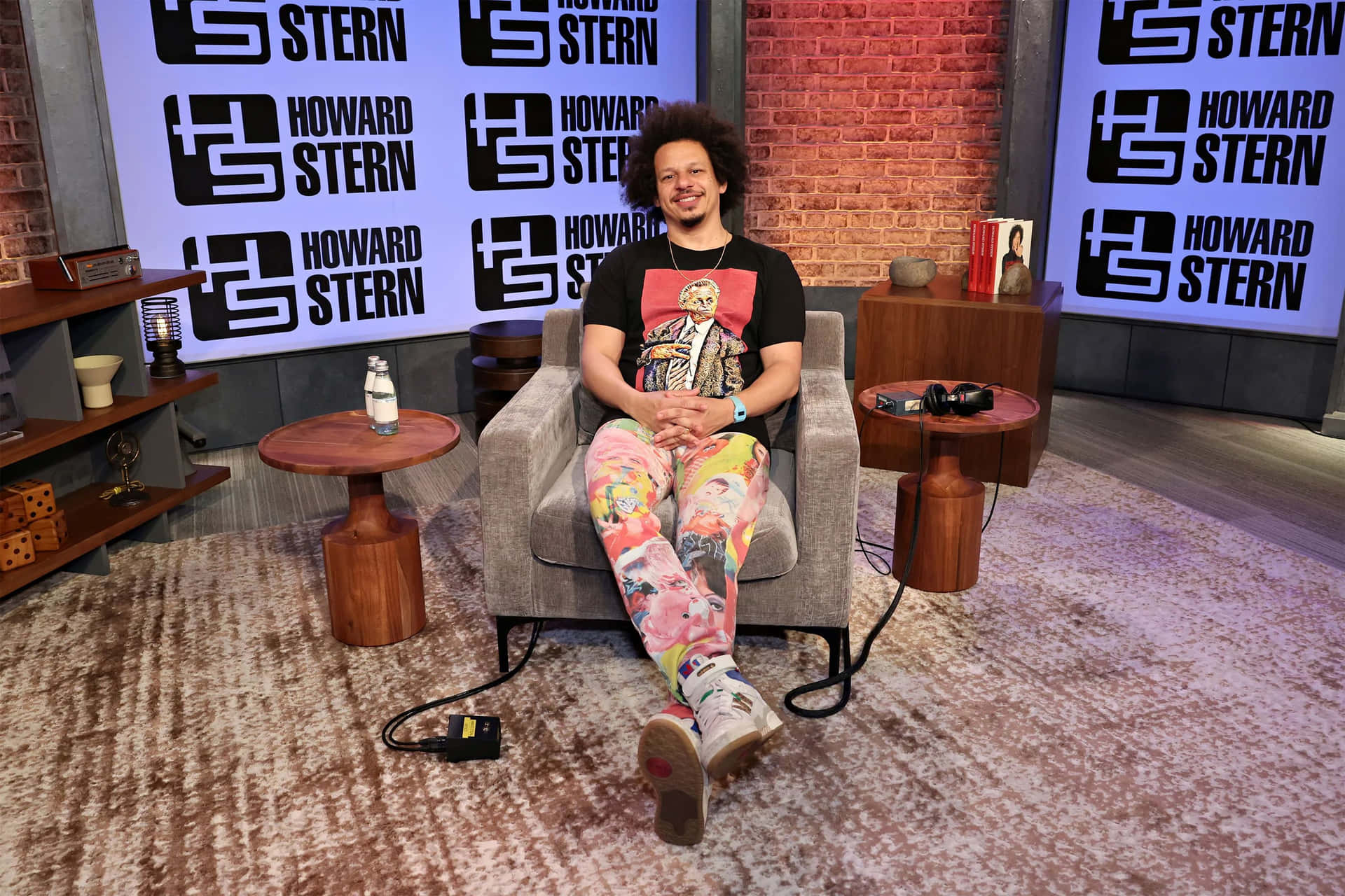  壁紙 — 一個捲髮男人放鬆地坐在灰色扶手椅上，身後是磚牆和'Howard Stern'標誌的脫口秀場景。