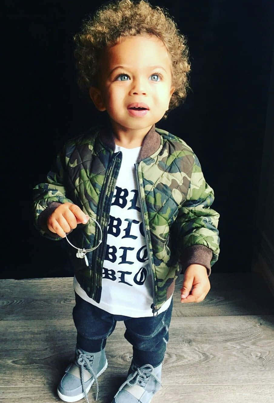  壁紙 — 一個捲髮幼兒穿著迷彩外套和圖案T恤，站在深色背景的木地板上。