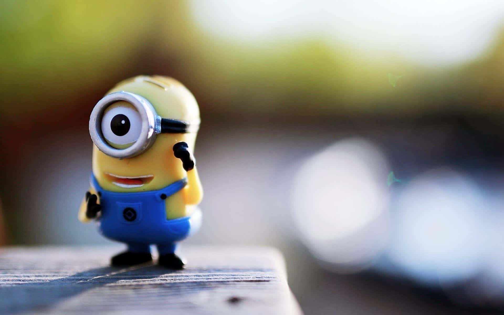  壁紙 — 一個開心的黃色Minion公仔，有一隻眼睛和藍色吊帶褲，站在木質表面上，背景是柔和模糊的戶外環境。