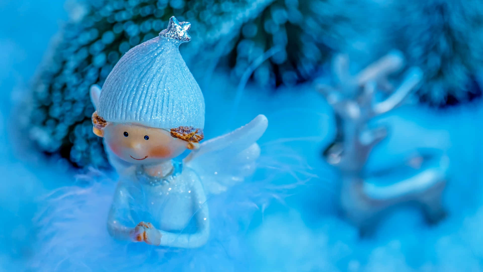 壁紙 — 一個快樂的雪天使公仔，戴著星形帽子，坐在雪白的藍色環境中，附近有一隻模糊的馴鹿。