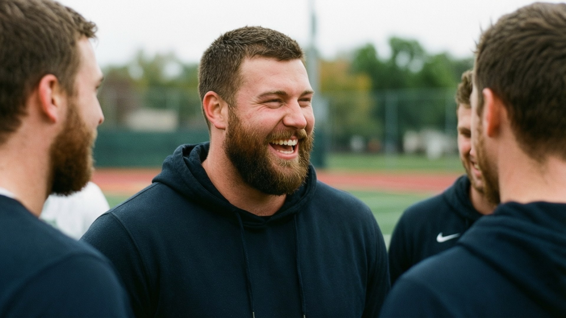 Teammate Laughter 壁紙 — 一個留鬍鬚的男人穿著深色連帽衫，在運動場上被隊友包圍，開懷大笑。