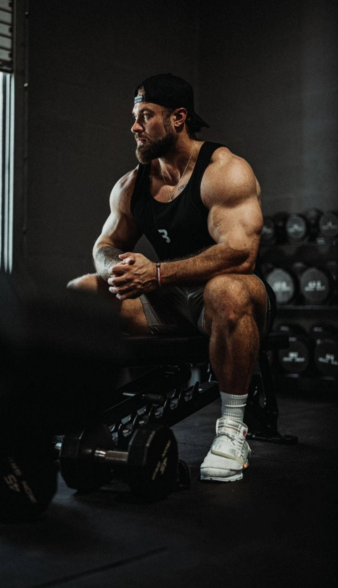 一個留鬍子的肌肉男坐在健身房的長凳上，周圍是槓鈴片。 — Chris Bumstead 壁紙