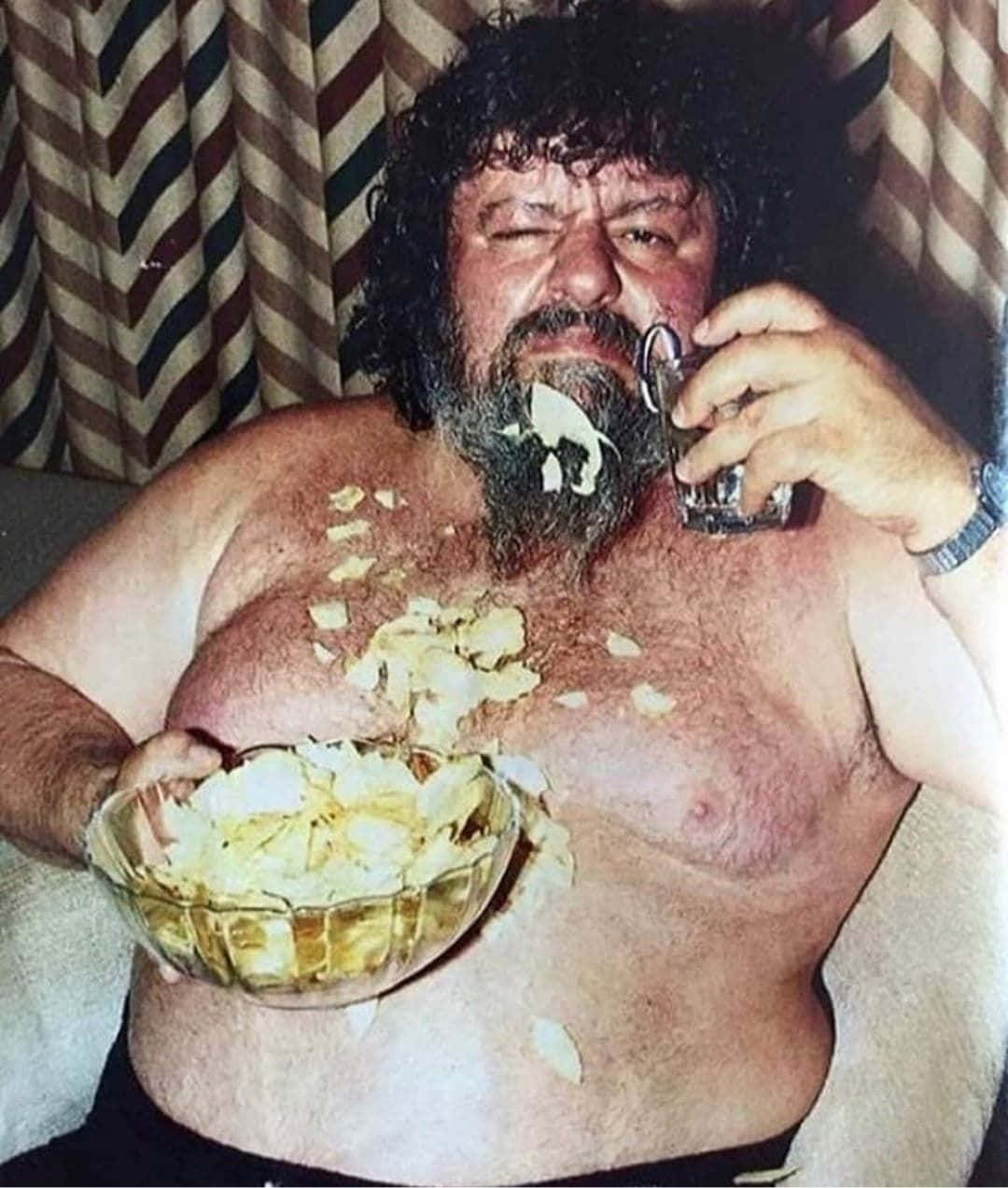  壁紙 — 一個沒穿上衣、留著鬍子和捲髮的男人坐在鋪滿洋芋片的沙發上，一邊喝著烈酒杯。