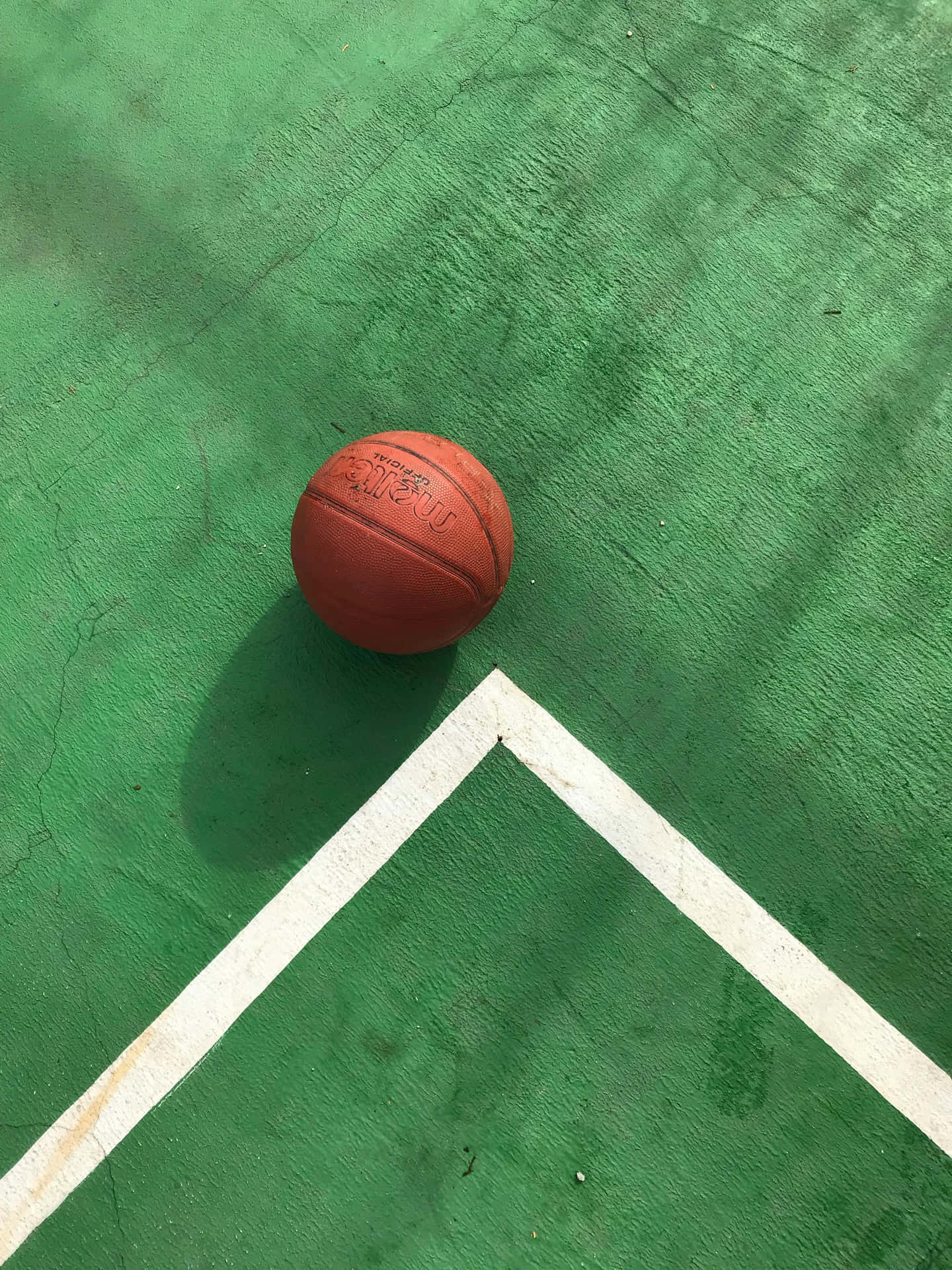  壁紙 — 一個磨損的橙色籃球放在有白色線條的裂開綠色球場上，在明亮的日光下。