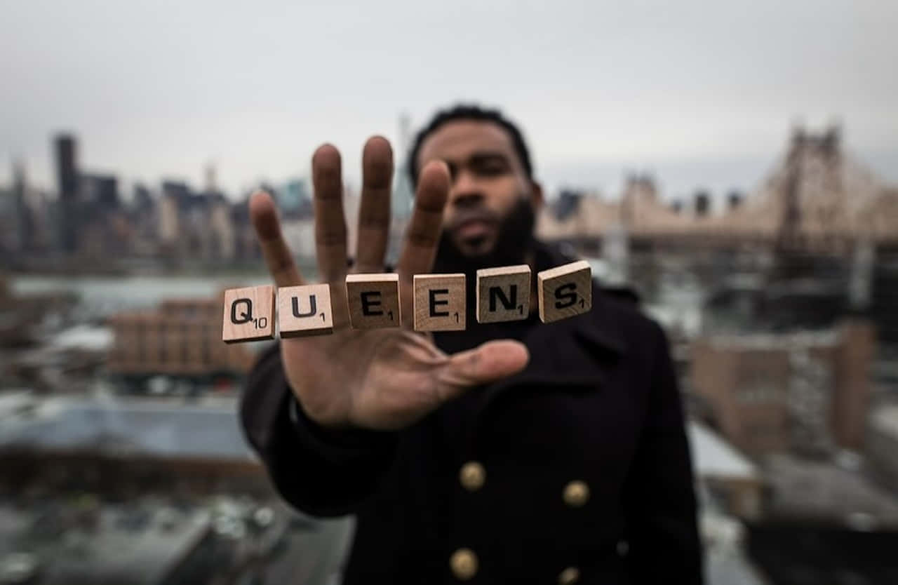  壁紙 — 一個男人拿著拼出 queens 的 Scrabble 字母牌，背景是模糊的紐約市天際線和橋樑。