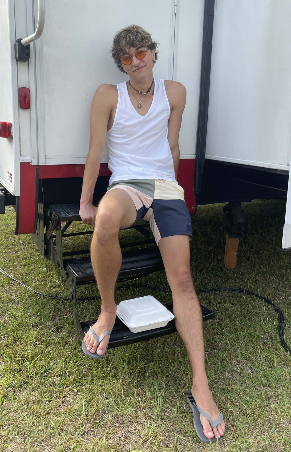 壁紙 — 一個年輕男子，捲髮，戴橙色太陽眼鏡，穿著白色背心和彩色短褲，悠閒地坐在白色拖車的台階上。